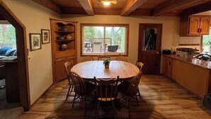 A wooden interior with a round dining table surrounded by chairs, a window with a view of a deck, and wooden cabinets.