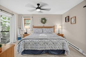 Ein Schlafzimmer mit Bett, Deckenventilator, Fenster mit Vorhängen, Nachttisch mit Lampe, Wandkrans und Blick ins Freie.