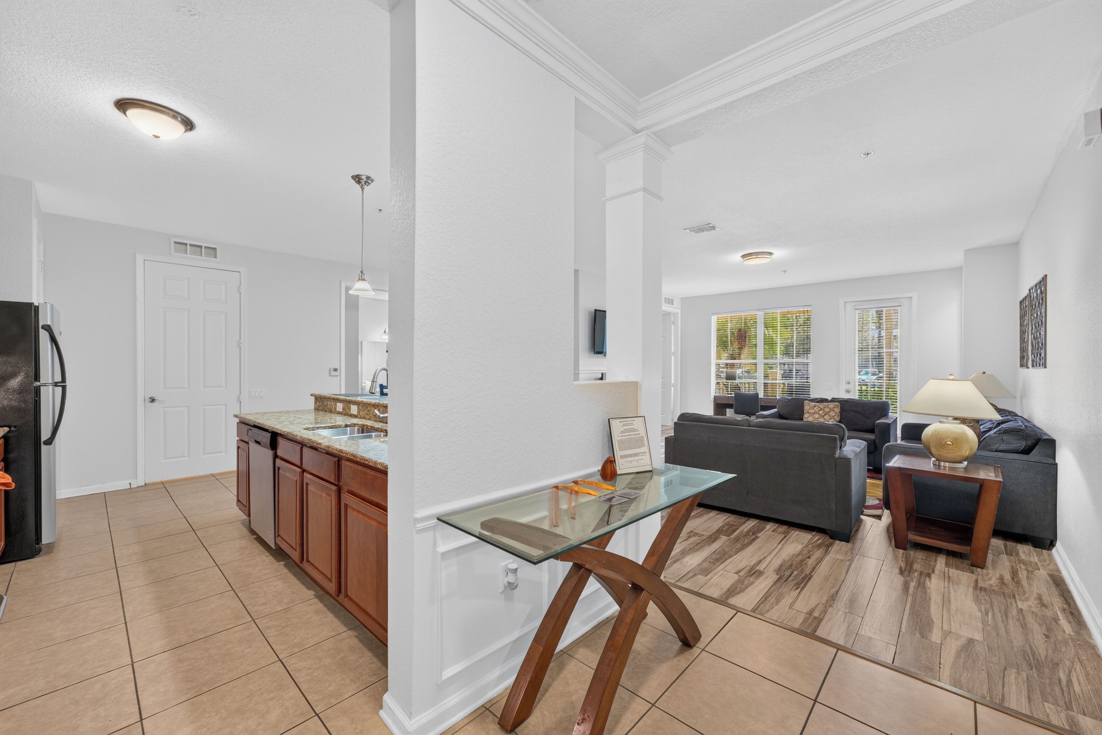 A modern kitchen with a built-in dishwasher, a dining area with a glass table, and a living room with a sofa and a lamp.