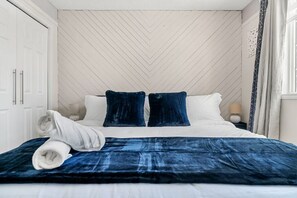 A neatly made bed with a blue blanket and pillows, a white headboard, and a white wall with a herringbone pattern.