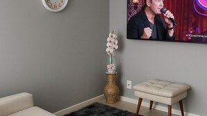 Een woonkamer met een bank, een klok, een vaas met bloemen, een bijzettafeltje en een televisie waarop een persoon te zien is.