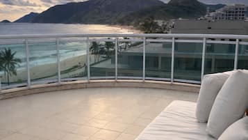 Un balcón con zona de estar con cojines blancos, baranda de vidrio y vista a la playa y las montañas.