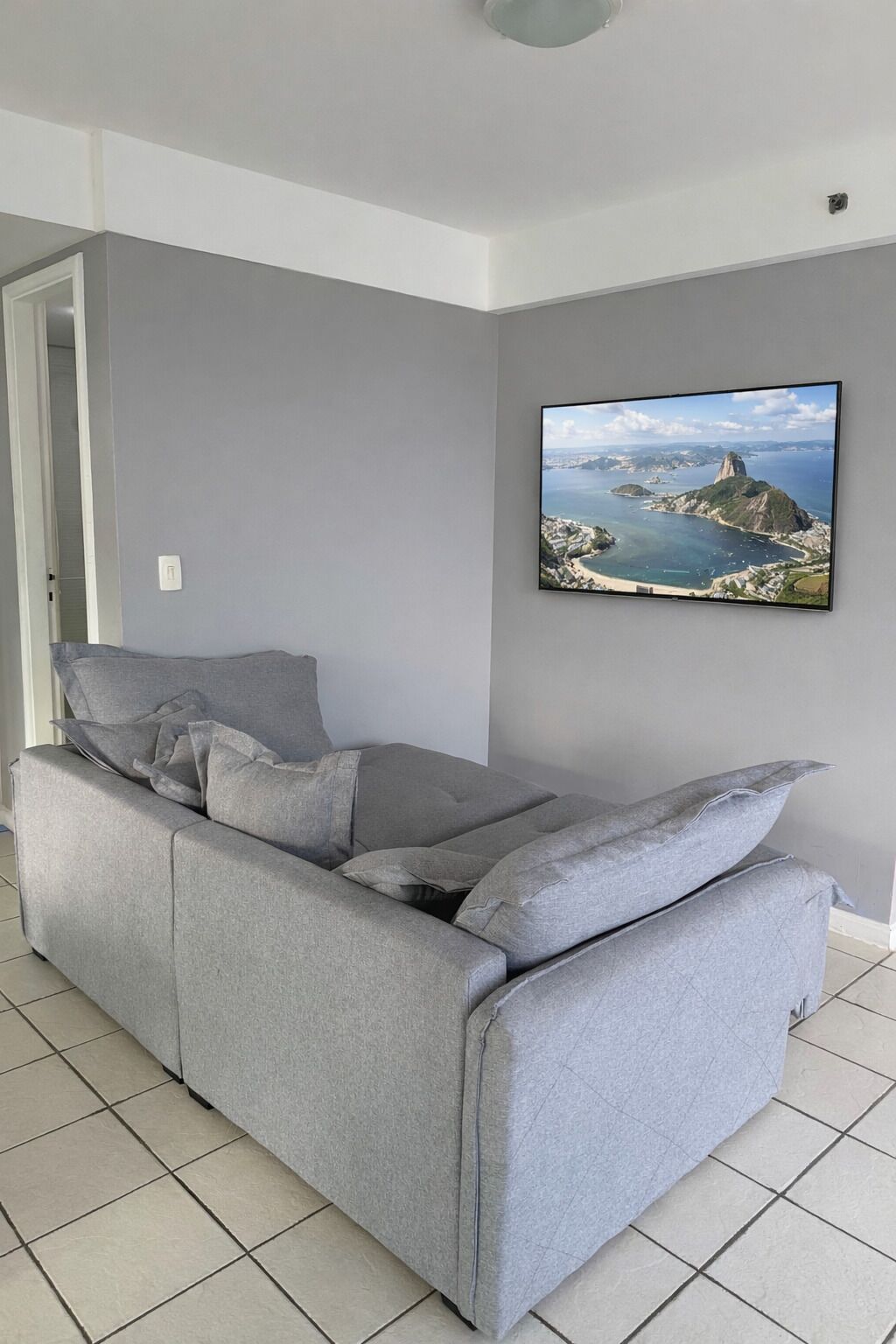 Un sofá gris en una habitación con suelo de baldosas y un cuadro en la pared que muestra un paisaje costero.