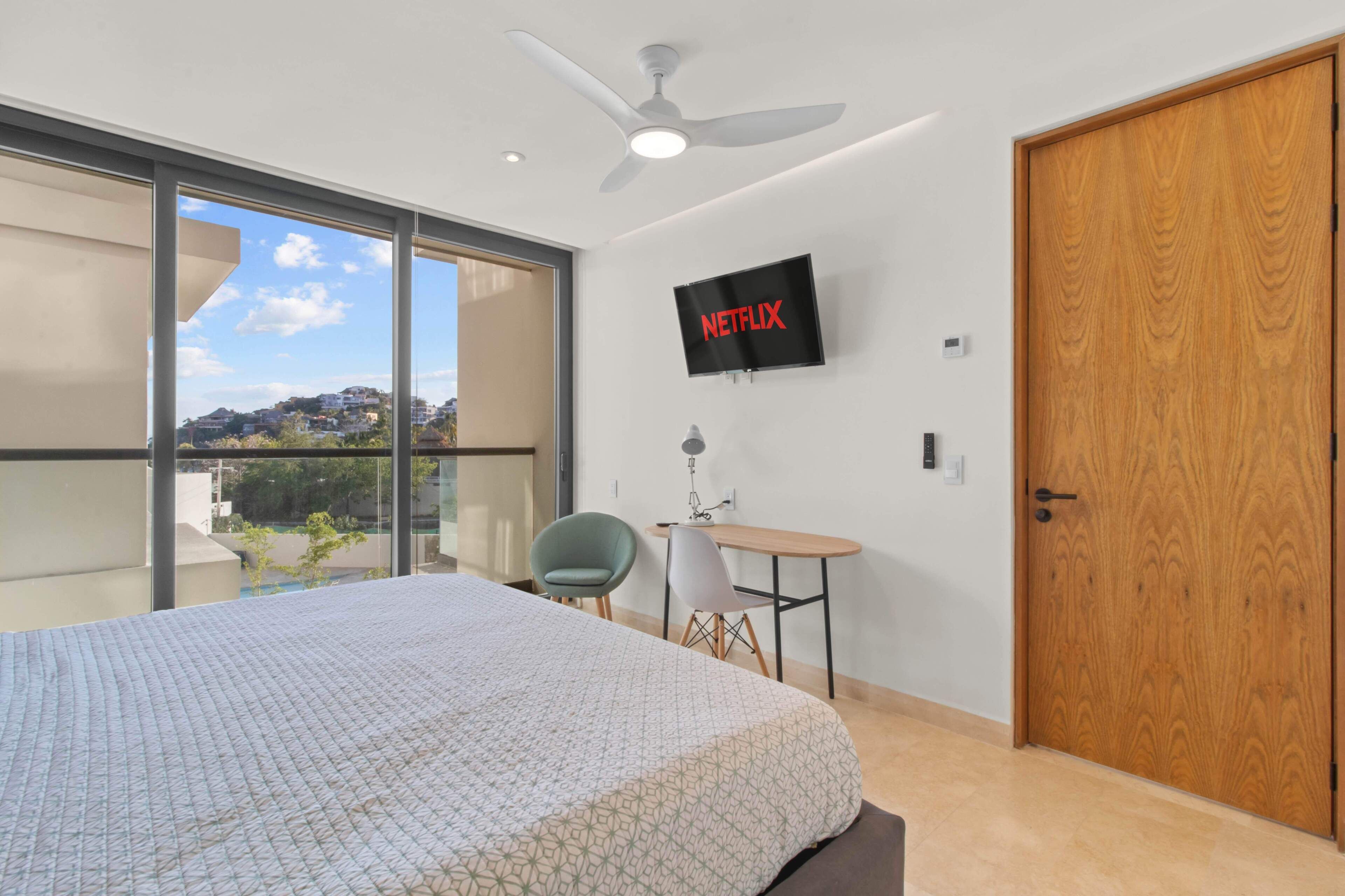 Un dormitorio moderno con una cama grande, un escritorio, una silla, un televisor montado en la pared y un ventanal corredero que da a un balcón.