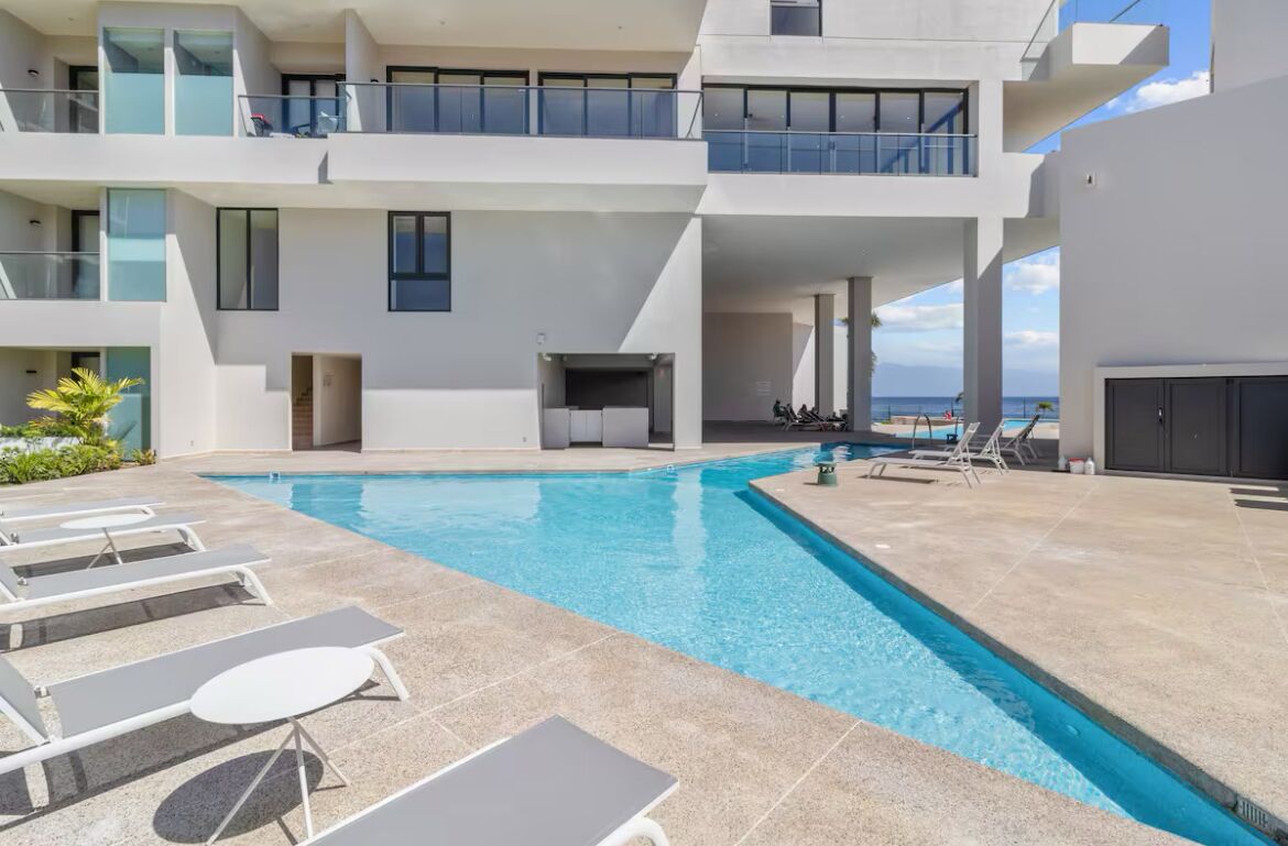 A modern hotel with a rooftop pool, sun loungers, and a view of the ocean.