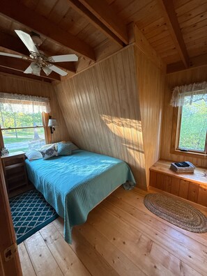 A wooden cabin bedroom with a bed, a ceiling fan, a window with curtains, a bedside table, and a rug.
