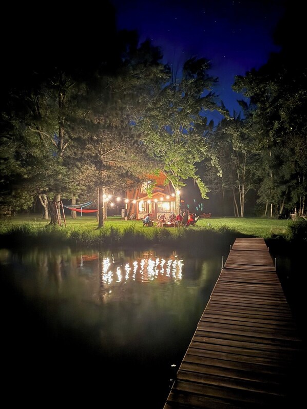A wooden dock leading to a house with string lights, surrounded by trees and a calm body of water.