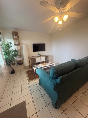 A living room with a green sofa, a flat-screen TV mounted on the wall, a ceiling fan, and a small table with a remote control. - The Perch on Hawkhill, Lower Level (Saint Thomas)