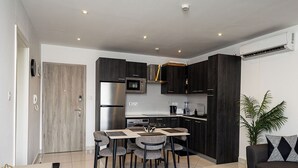 A modern kitchen with dark cabinetry, a stainless steel refrigerator, and a microwave. There is a dining area with a table and chairs. - Cozy One bed at Loxwood (Accra)