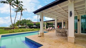 Zona de piscina techada al aire libre con sillones de descanso y una mesa, rodeada de palmeras y un jardín ajardinado.