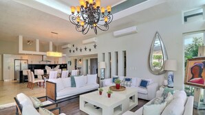Amplia sala de estar con un sofá blanco, una mesa de centro y un candelabro. Se observa una cocina al fondo con electrodomésticos modernos y un comedor con sillas y una mesa.
