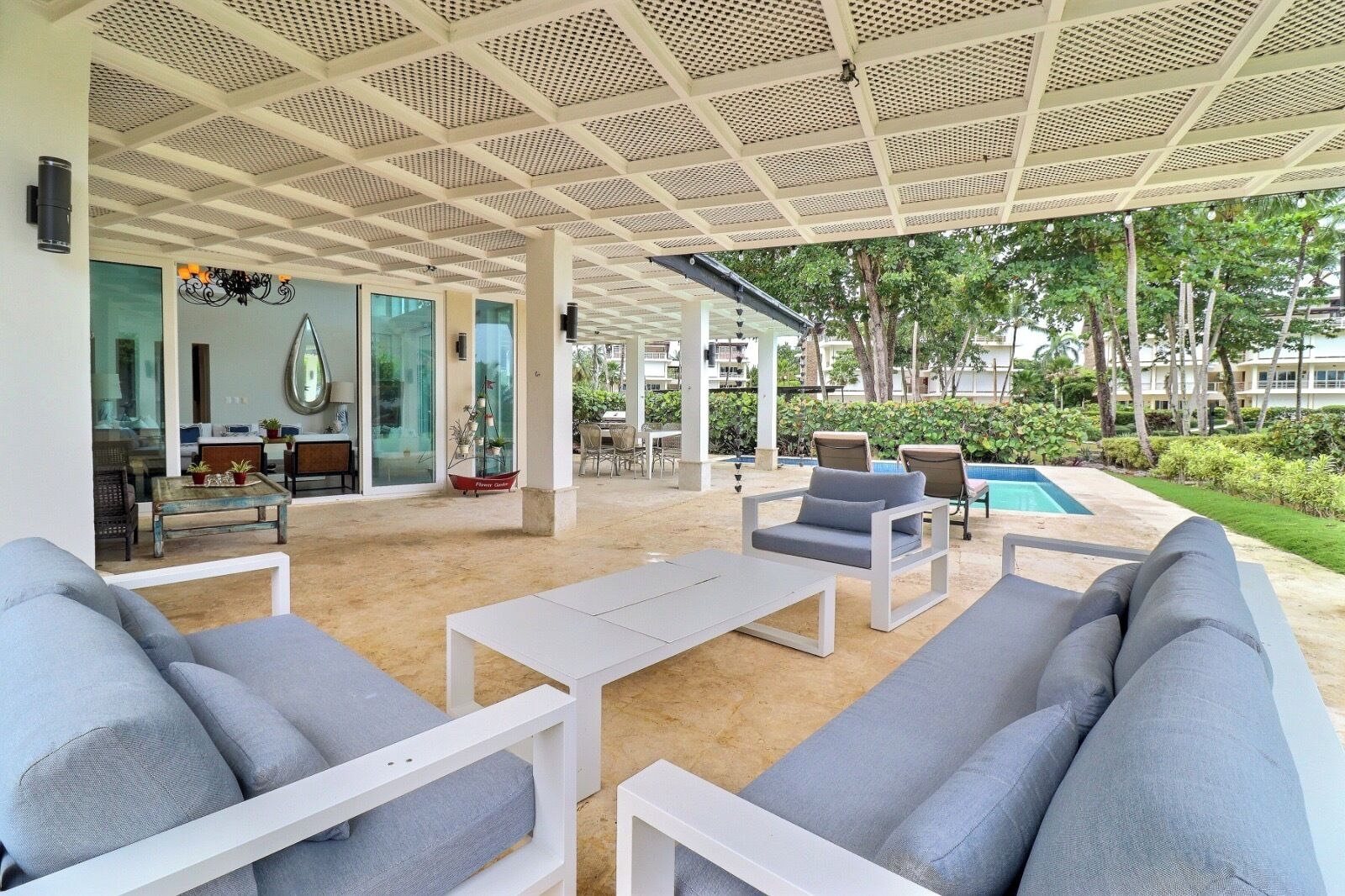 A covered outdoor seating area with modern furniture, a pool, and lush greenery.