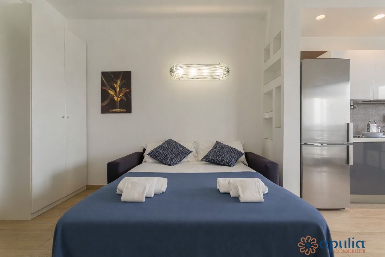 Ein modernes Hotelzimmer mit Bett, Kissen, einer Wandleuchte, einem Gemälde und einer Küchenzeile.