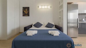 A modern hotel room with a bed, pillows, a wall-mounted light, a painting, and a kitchen area. - Welcome to Bari Sky Terrace (Bari)