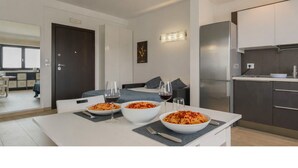 A modern kitchen with white cabinets, a stainless steel refrigerator, and a dining area with a white table set for two with pasta dishes and wine glasses. - Welcome to Bari Sky Terrace (Bari)