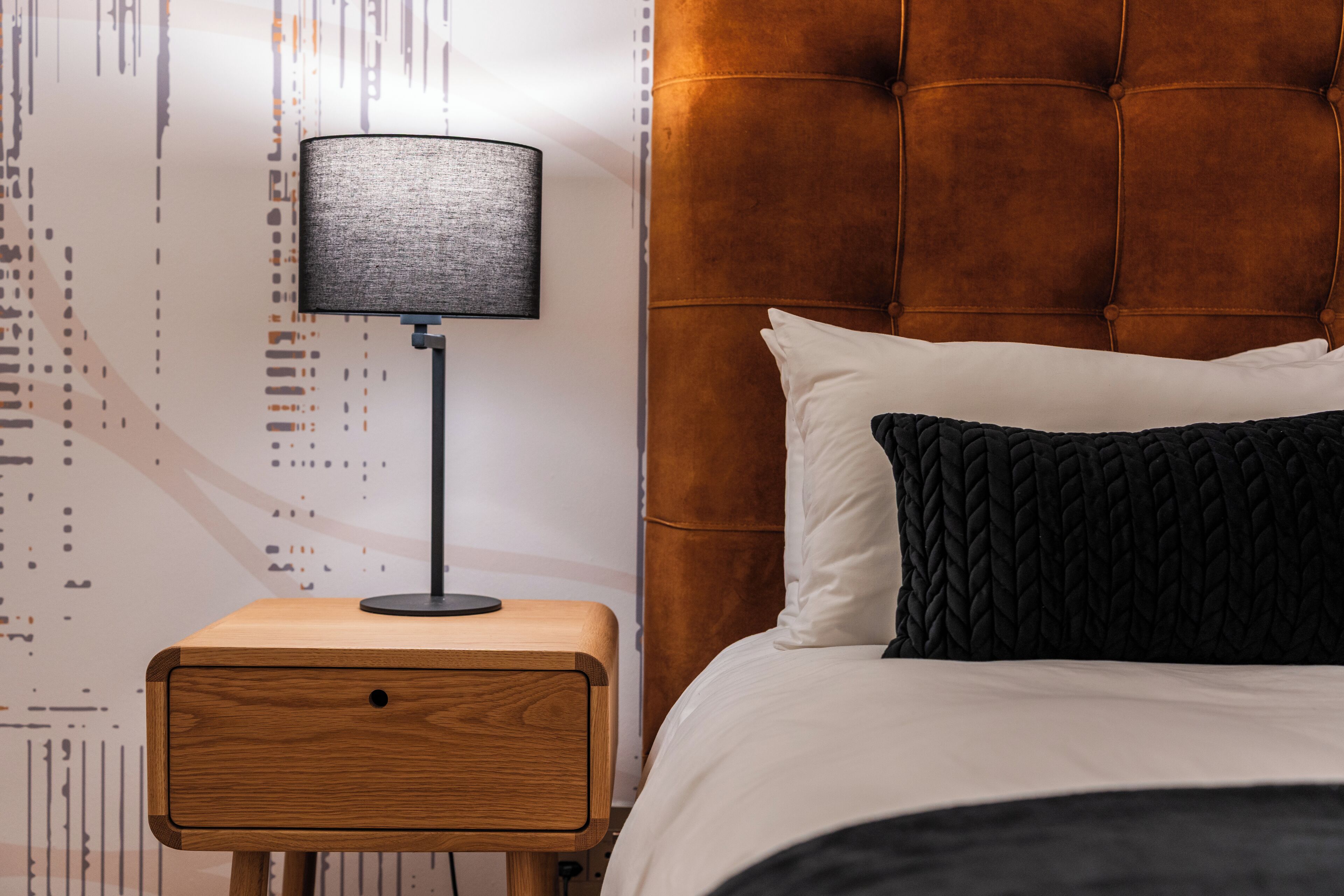 A bedroom with a wooden nightstand, a black lamp, a tufted headboard, and a bed with white and black bedding.