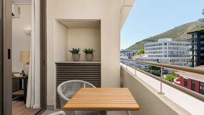 A balcony with a wooden table and wicker chair, two potted plants, and a view of buildings and a hill. - Dolce Vita | Lush Sea Point Gem with Balcony (Cape Town)