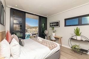 Un dormitorio moderno con cama, escritorio, silla y vistas a un paisaje urbano a través de amplios ventanales.