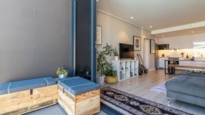 En moderne stue med sofa, træbænk og TV. I baggrunden ses et køkken med skabe og spiseplads.