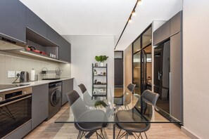 A modern kitchen with grey cabinets, a built-in oven, and a washing machine. There is a glass dining table with chairs and a small potted plant on a shelf. - The Lineal | Luxury Living for Work and Leisure (Johannesburg)