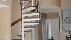 Un escalier en colimaçon avec une rampe en bois et des marches blanches, menant à un palier où se trouvent une table à manger et des chaises.