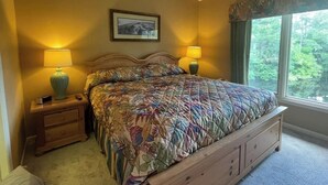 Une chambre avec un grand lit, deux lampes de chevet, une table de nuit, un tableau encadré au mur et une fenêtre donnant sur un paysage verdoyant.