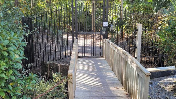 Une promenade en bois mène à une entrée fermée par un portail, qui est elle-même entourée d’une végétation luxuriante.