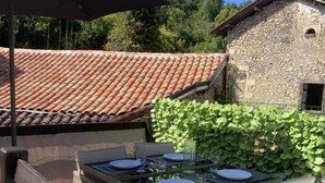 Un espace de restauration en plein air avec une table dressée pour quatre personnes, entouré de chaises en osier, un bâtiment en pierre avec un toit en tuiles et un mur recouvert de lierre.