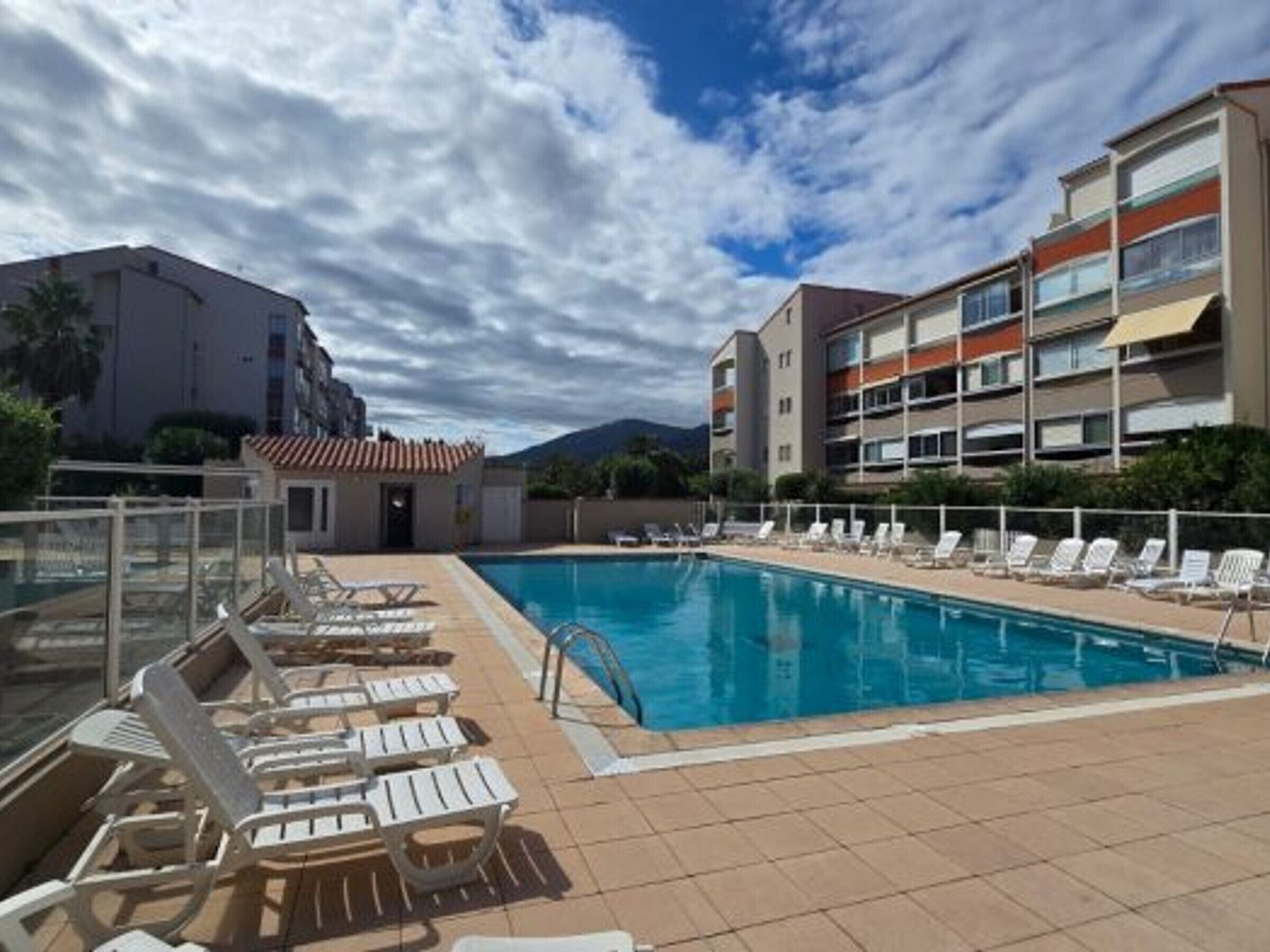 A swimming pool with lounge chairs, a glass balustrade, and a multi-story building in the background.
