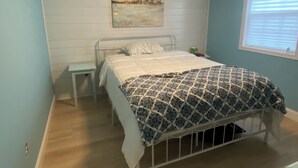 A bedroom with a bed, a nightstand, a ceiling fan, a window with blinds, and a wall-mounted air conditioner. -  Campfire Bliss at the Beach 
Eastern Shore Virginia Cottage Getaway (Horntown)