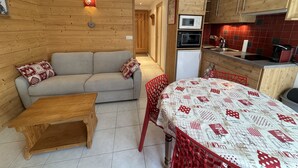 A compact living space with a sofa, a small wooden table, a dining table with red chairs, a kitchen area with wooden cabinets, a microwave, and a refrigerator. - Studio cabin in the center - 4 guests (Les Carroz d'Arâches)