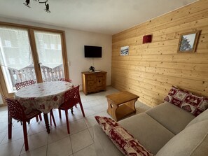 Ein Wohnzimmer mit einer Couch, einem Esstisch mit Stühlen, einem Holzschrank und einem an der Wand befestigten Fernseher.