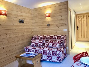 A cozy room with a wooden wall, a patterned sofa, a small table with a magazine, and a red chair. - Studio cabin in the center - 4 guests (Les Carroz d'Arâches)
