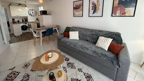 Una sala de estar moderna con un sofá gris, una mesa de centro y un comedor con una mesa y sillas. Se observa una cocina al fondo con una lavadora y un microondas.