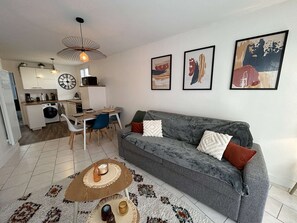 Un salon moderne avec un canapé gris, une table basse et un coin repas comprenant une table et des chaises. On aperçoit une cuisine au fond, avec un lave-linge et un four à micro-ondes.