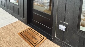 Eine schwarze Tür mit Glasfenster, eine Fußmatte mit der Aufschrift "WELCOME" und zwei Wandleuchten.