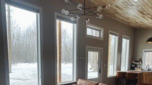 Ein geräumiges Wohnzimmer mit einer Ledersofa, einem runden Couchtisch und einer Deckenleuchte. Große Fenster bieten Blick auf eine verschneite Landschaft.