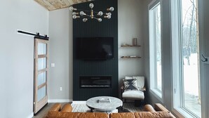 Ein modernes Wohnzimmer mit Holzboden, einem großen braunen Ecksofa, einem runden Couchtisch, einem an einer dunklen Wand montierten Fernseher und großen Fenstern mit Blick auf Bäume.