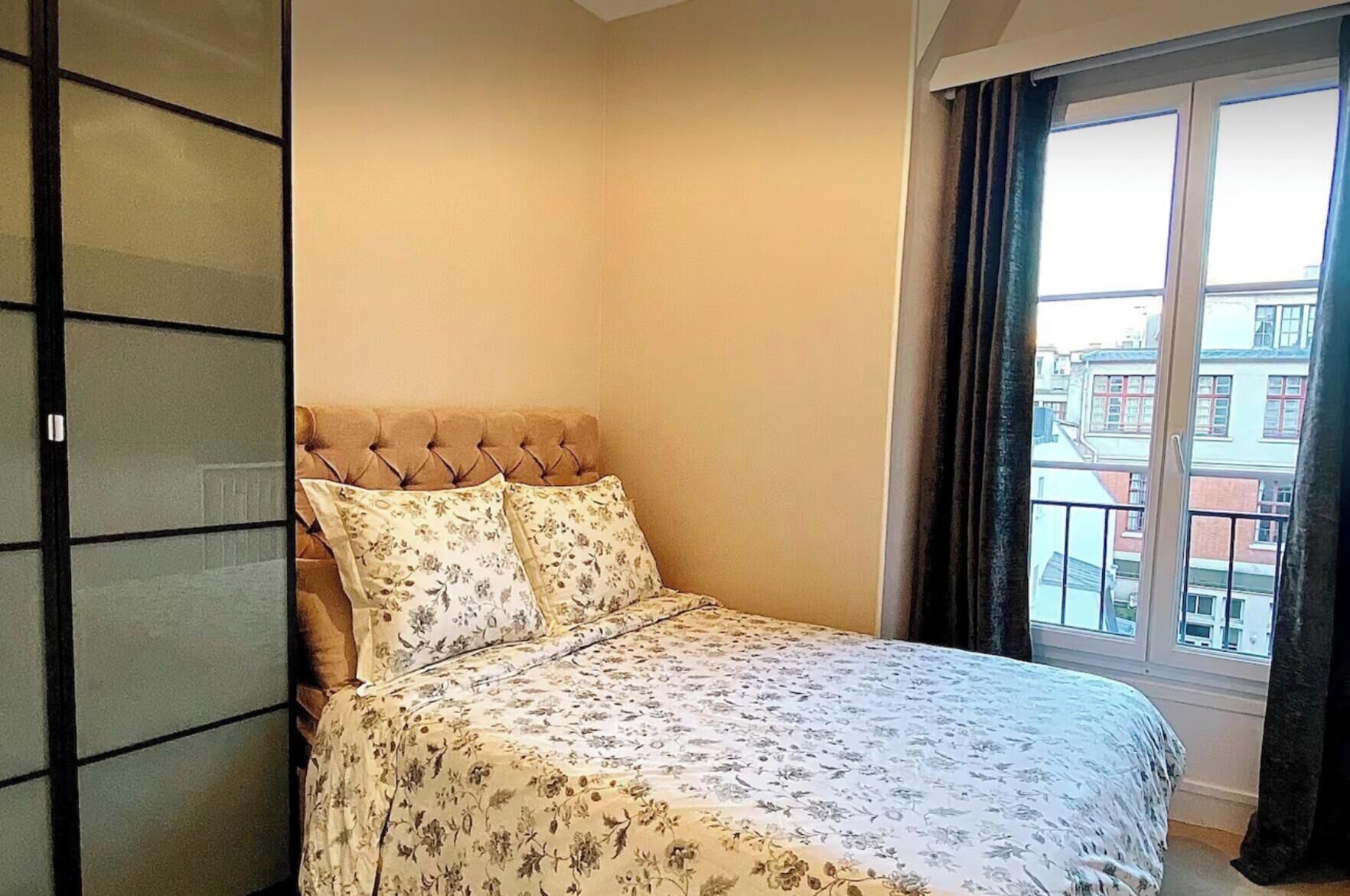 A bedroom with a bed, a window, and a view of buildings outside.
