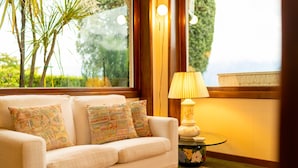 Ein gemütliches Wohnzimmer mit einer Couch, gemusterten Kissen, einem kleinen Tisch mit einer Lampe und Blick auf Grünflächen draußen.