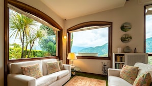 Ein gemütliches Wohnzimmer mit zwei weißen Sofas, einem kleinen Beistelltisch und einer Lampe. Große Fenster bieten Blick auf Grünflächen und Berge.