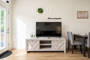 A living room with a TV on a wooden TV stand, a dining table with chairs, and a 'HOME' sign on the wall. - Elegant New-Build Suite • Walk to Everything (Hot Springs)