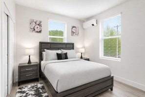 Une chambre à coucher comprenant un lit, deux tables de chevet, une commode, un climatiseur fixé au mur et deux fenêtres équipées de stores.