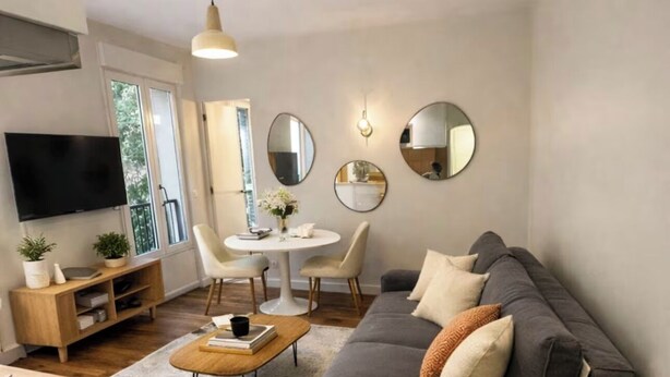 Een moderne woonkamer met een grijze bank, een salontafel en een televisie. Er is een eethoek met een witte tafel en stoelen.
