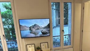 Een moderne woonkamer met een grote televisie, houten meubels en grote ramen met uitzicht op bomen en een gebouw.