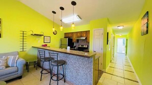 Cozinha compacta com bancada, mesa de jantar com cadeiras e sofá em um quarto amarelo e luminoso.