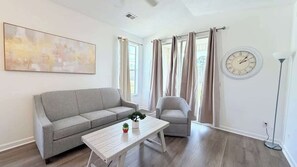 A modern living room with a grey sofa, a white coffee table, a small plant, and a clock on the wall. - Sunny River Oaks Retreat  2BR Golf Condo (Myrtle Beach)