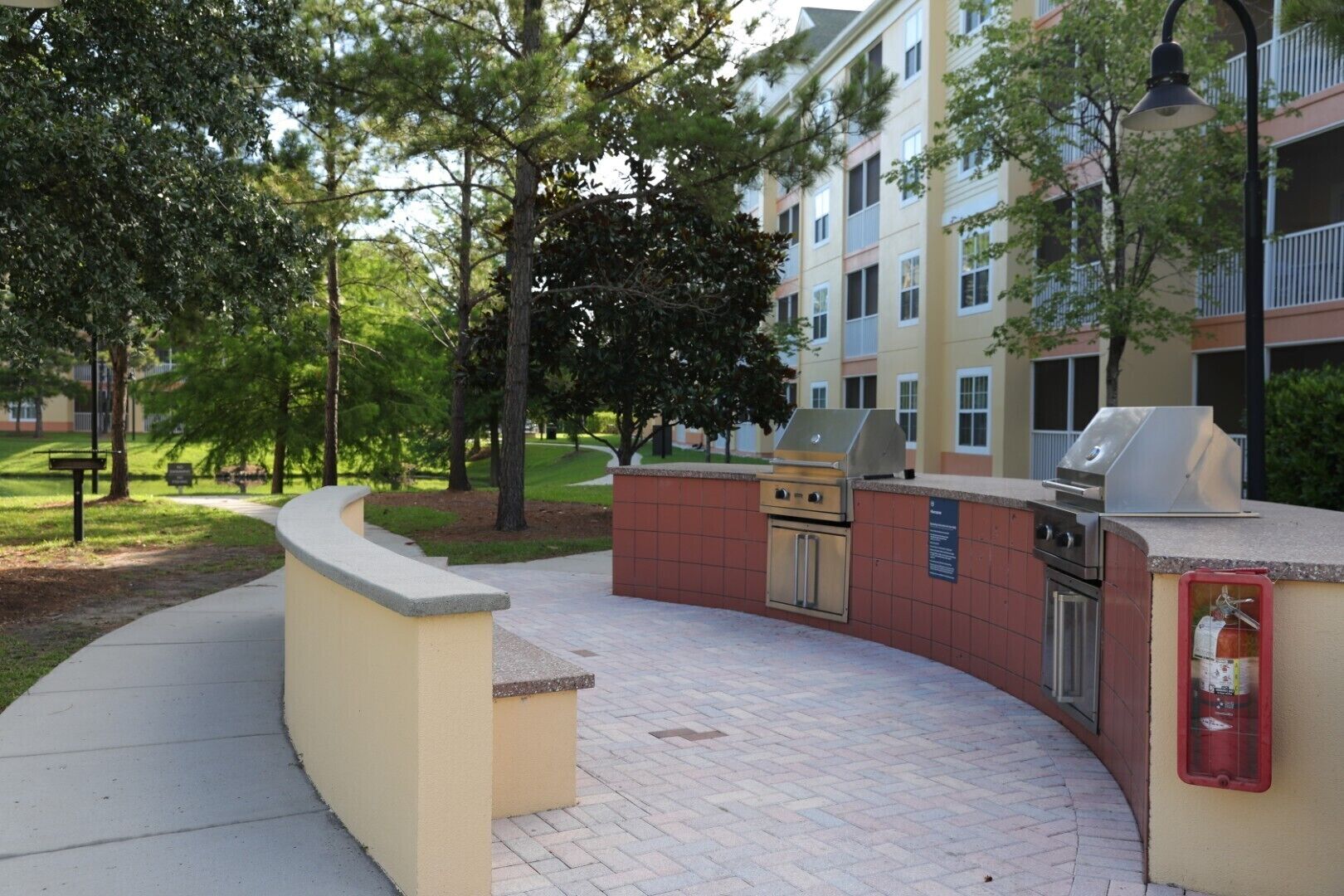 An outdoor barbecue area with a grill and a fire extinguisher, surrounded by a brick pathway and trees.