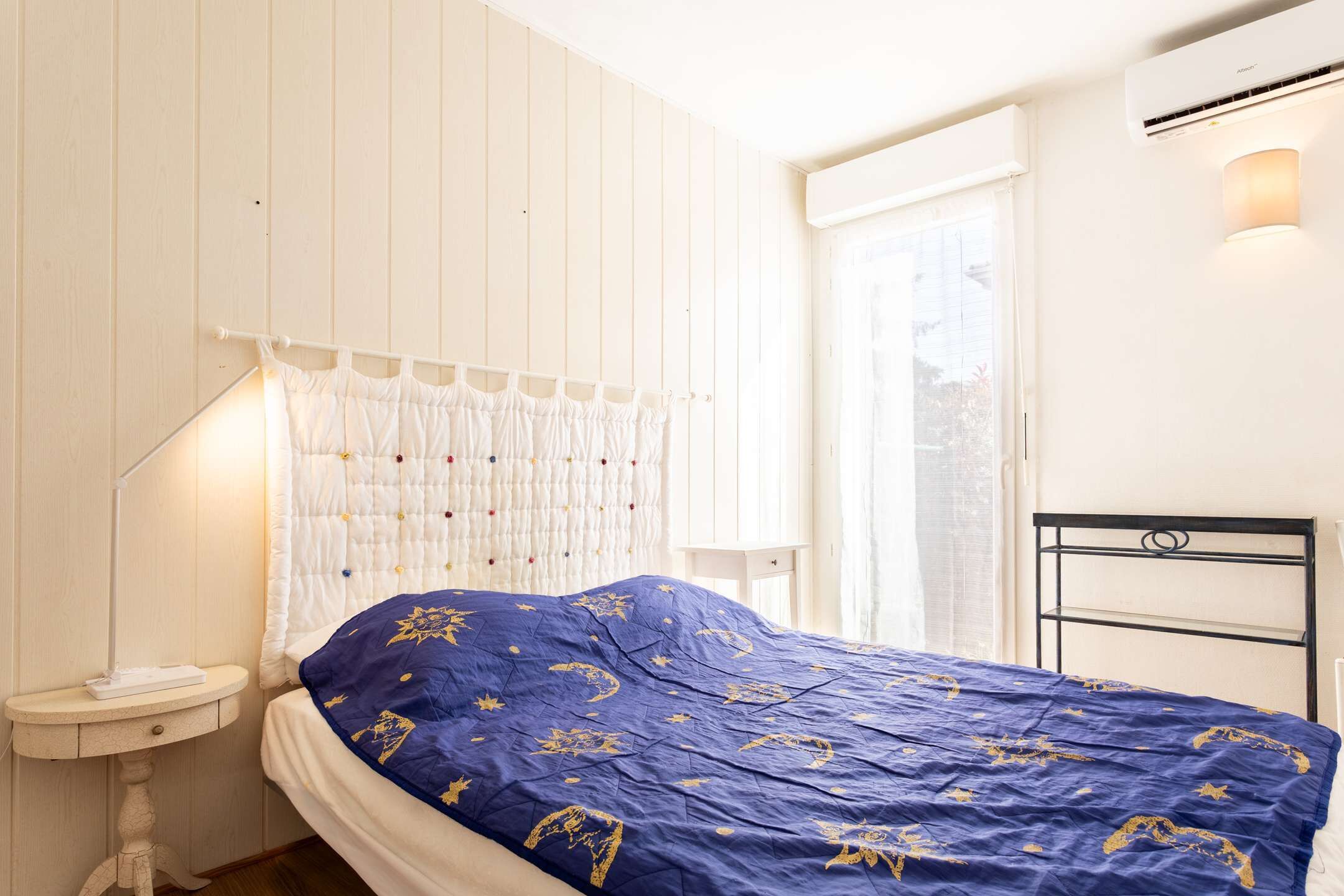 Una cama con colcha azul, una mesita de noche con lámpara, aire acondicionado instalado en la pared y una ventana con cortinas.