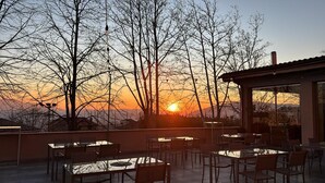 Un área de estar al aire libre con mesas y sillas, con vista a un atardecer con árboles desnudos y una cadena montañosa distante.